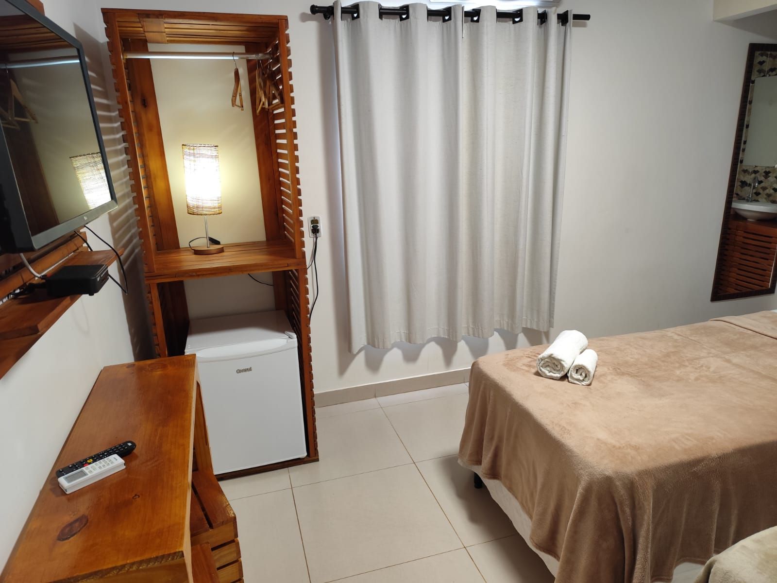 Quarto de hotel com cama, guarda-roupa, frigobar, televisão e janela com cortinas.