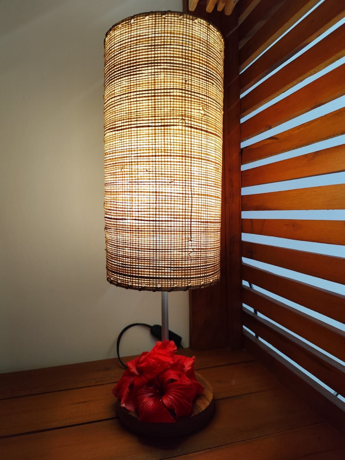 Abajur cilíndrico alto com cúpula trançada sobre superfície de madeira, ao lado de um arranjo de flores vermelhas.