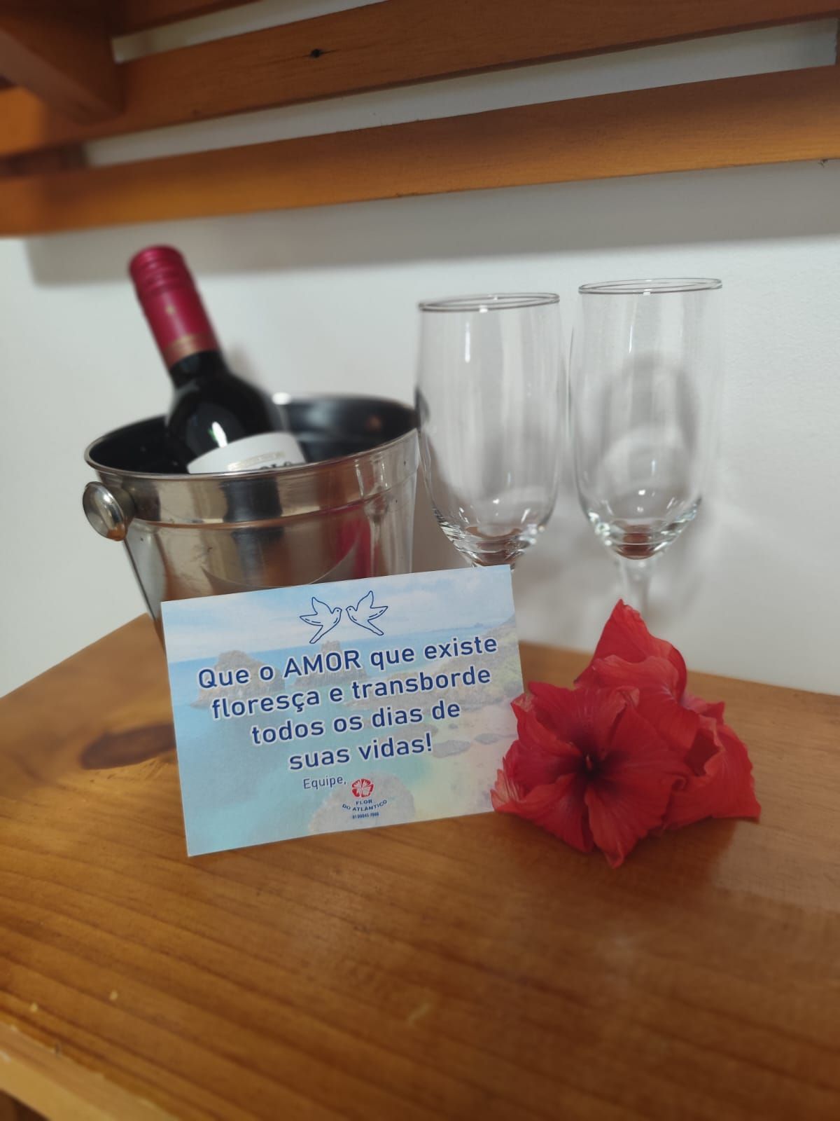 Garrafa de vinho em balde com taças, cartão e flor sobre superfície de madeira.