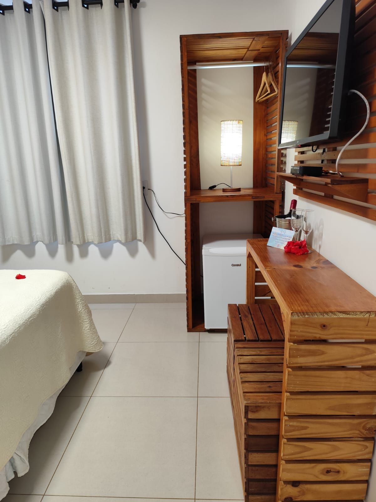 Quarto com móveis de madeira, TV, frigobar e cortinas brancas.