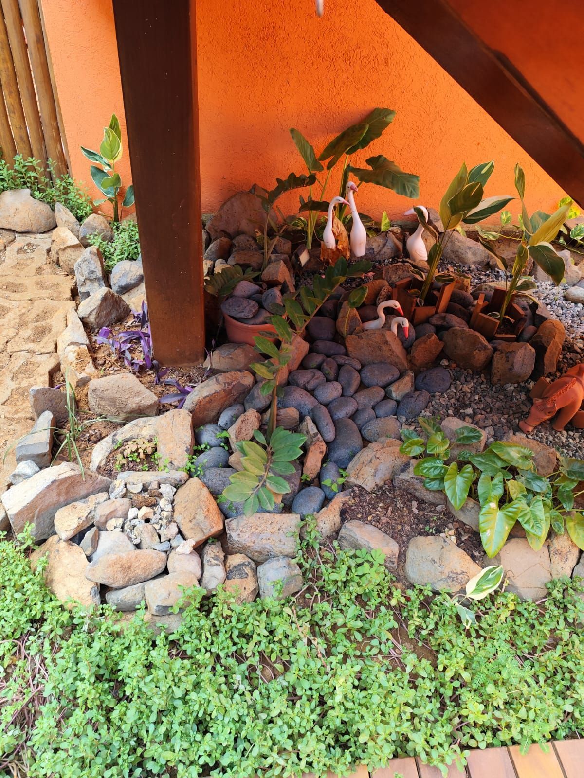 Canteiro de jardim com tijolos, pedras, plantas e estatuetas perto de uma viga de sustentação marrom contra uma parede laranja.