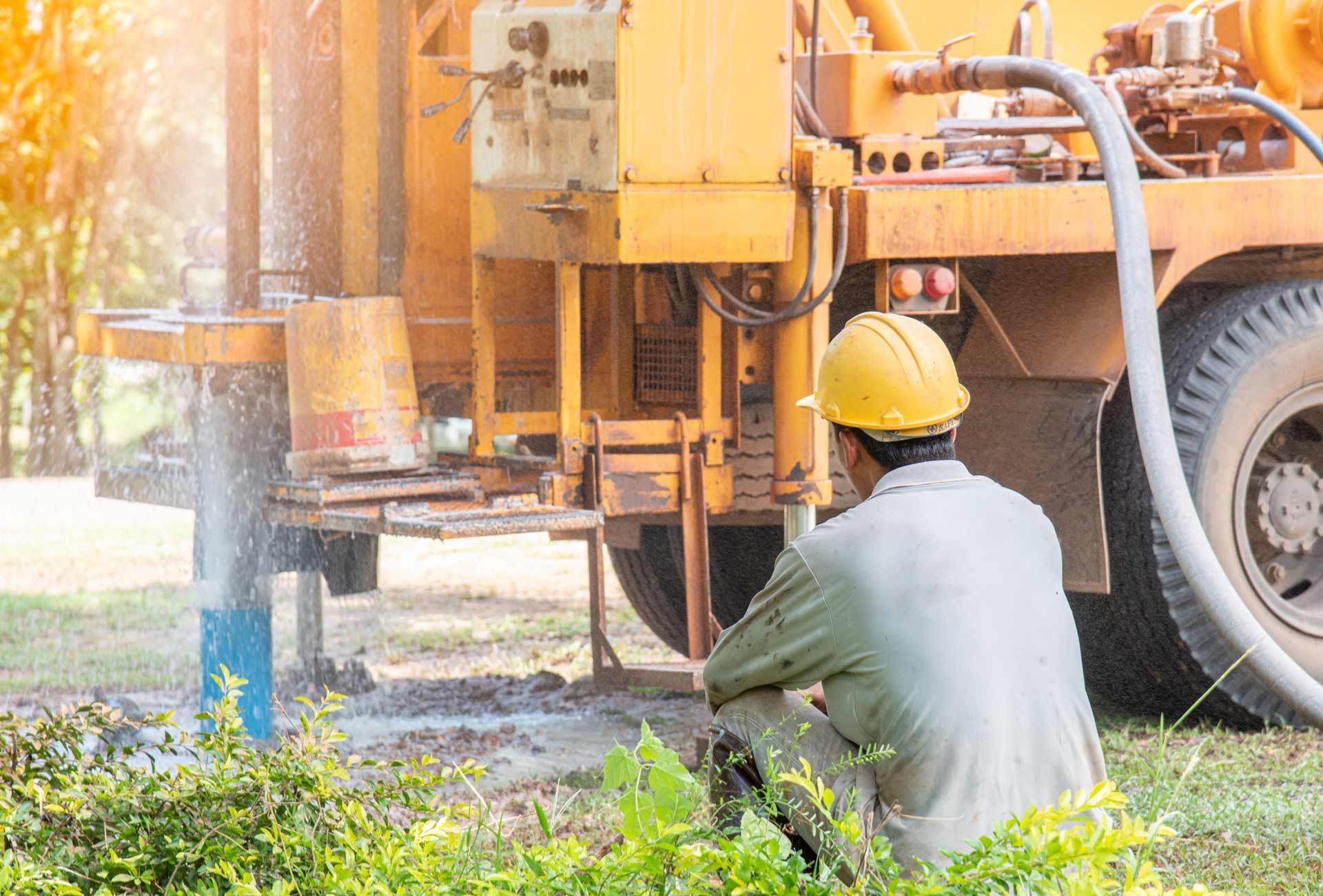Man in hard hat watches water erupt from well drilling site, near yellow machinery