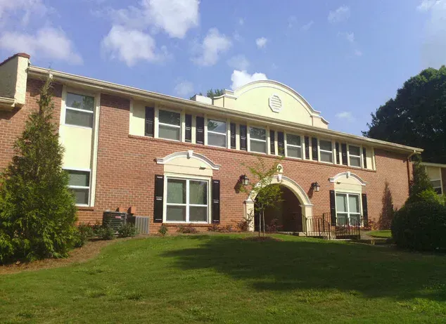 Two-story brick building with multiple windows, black shutters, and an arched entrance, set on a green lawn under a blue sky.