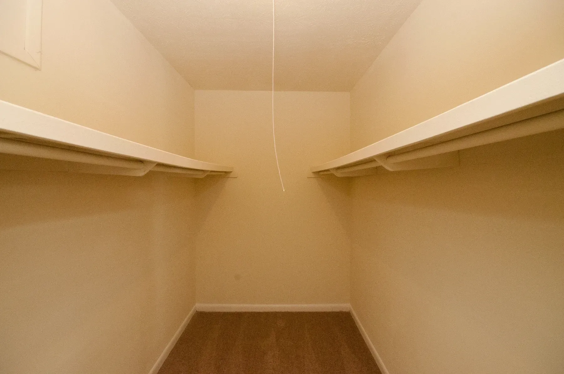 Empty walk-in closet with beige walls, two shelves, and brown carpet. A light cord hangs from the ceiling.