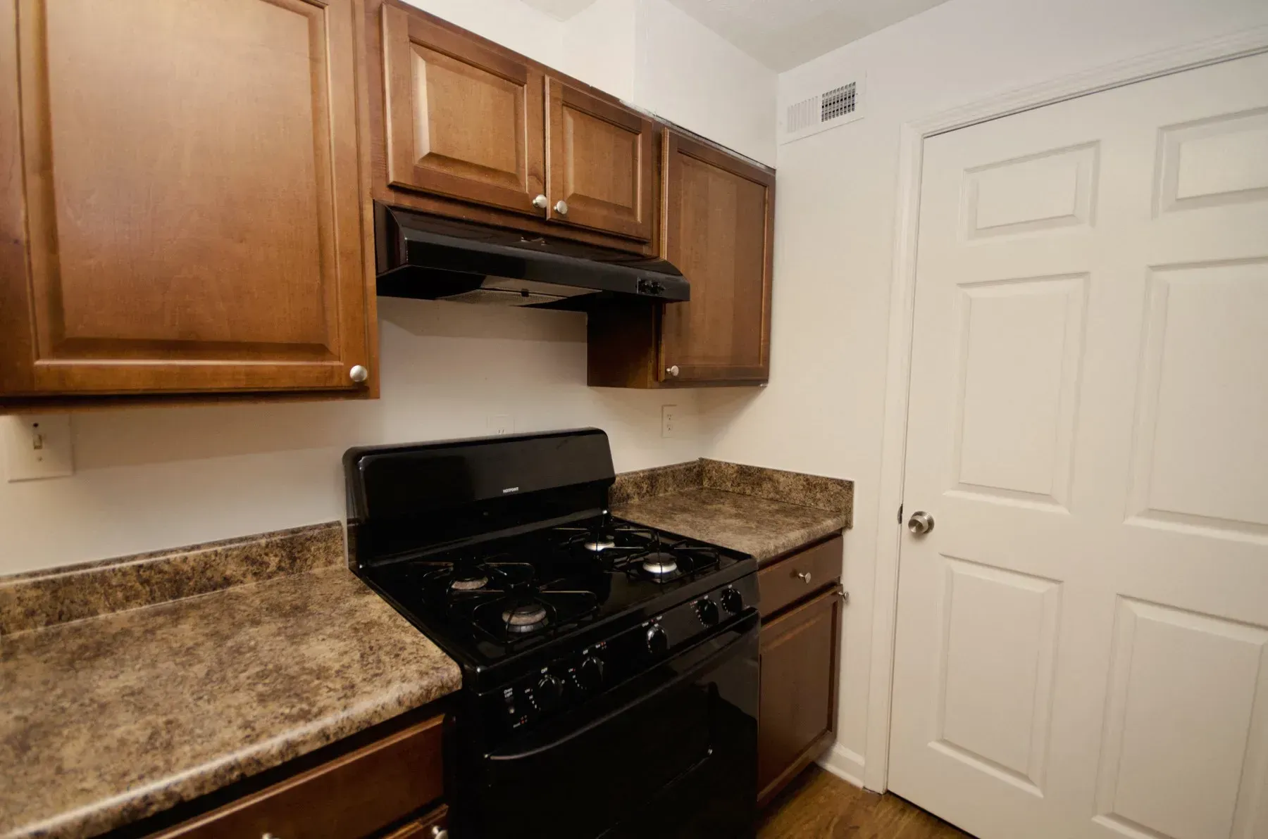 Kitchen with dark brown cabinets, black stove, and white door. Brown speckled countertops.