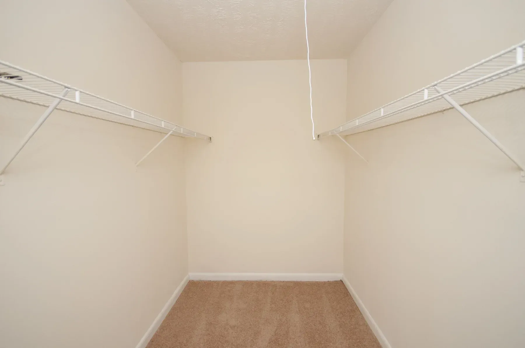 Empty walk-in closet with white wire shelving and tan carpet. Light tan walls and white ceiling.