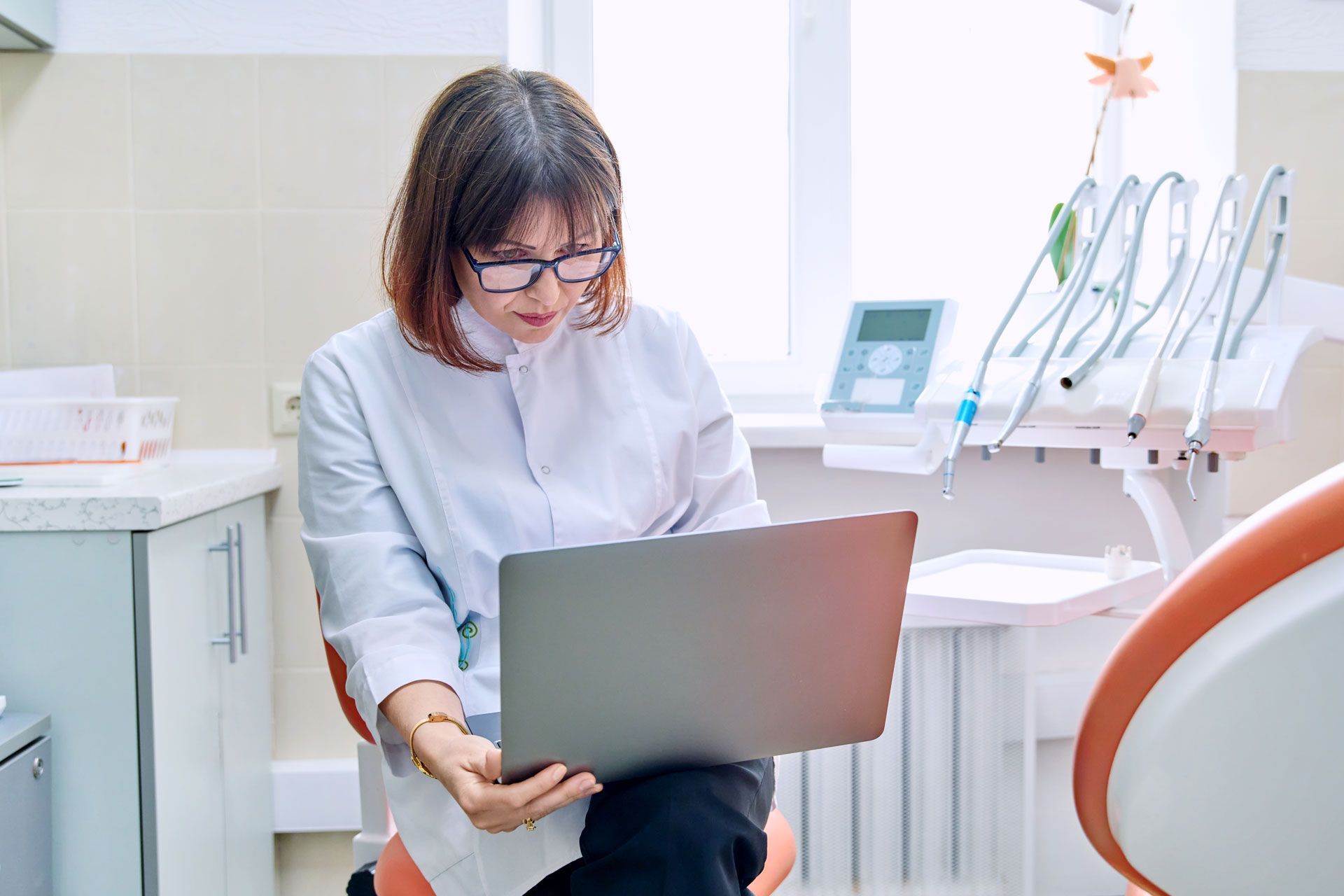 Dentist in white coat sits in dental chair, looking at a laptop in a dental office.