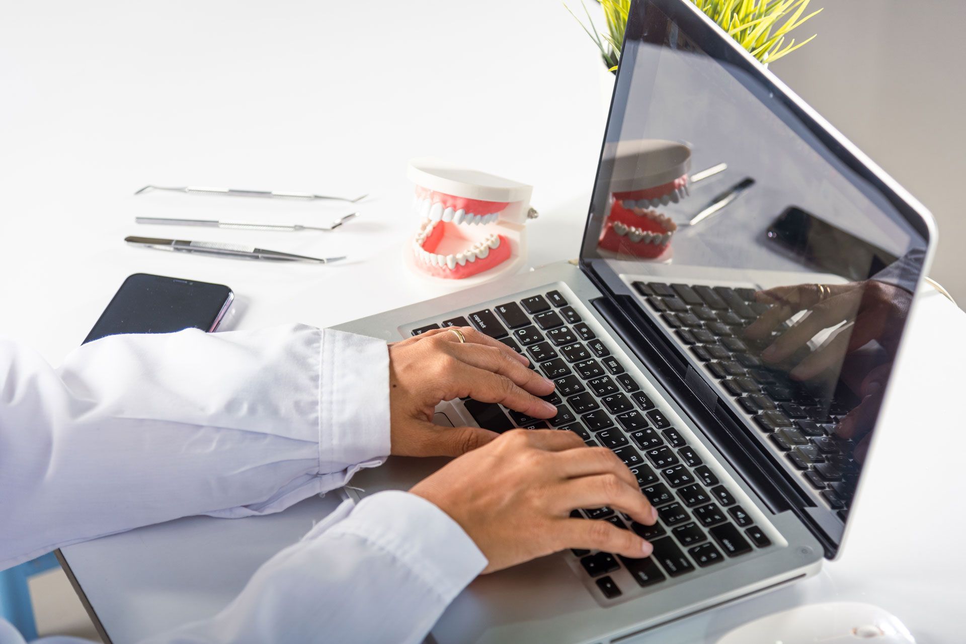 Hands typing on laptop at a dentist's office desk with dental tools, models, and a phone.