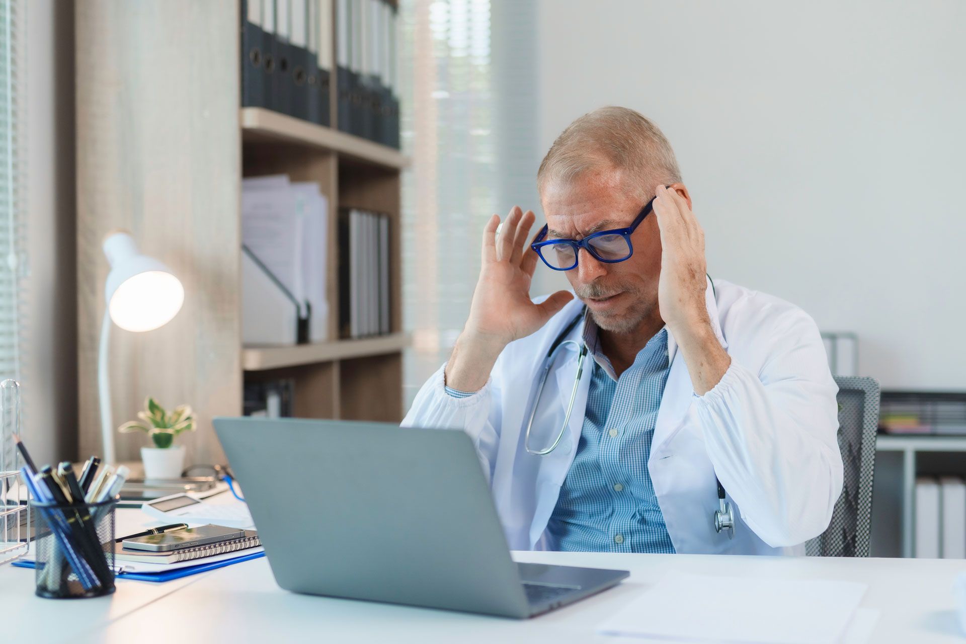 Doctor wearing glasses, in lab coat, holding head, looking at laptop in office.