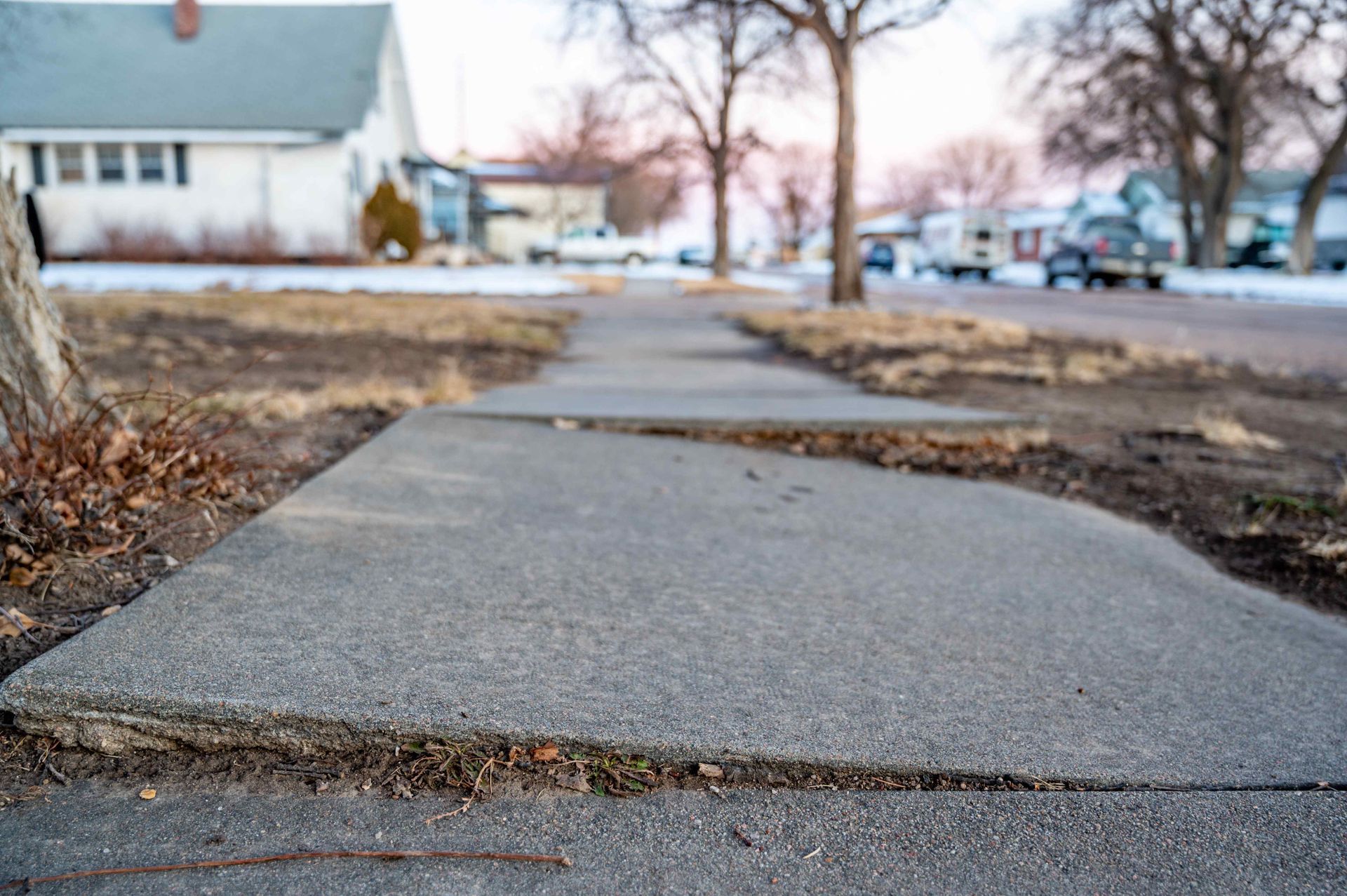 A residential sidewalk with a large, uneven gap and displacement between concrete slabs, creating a tripping hazard.