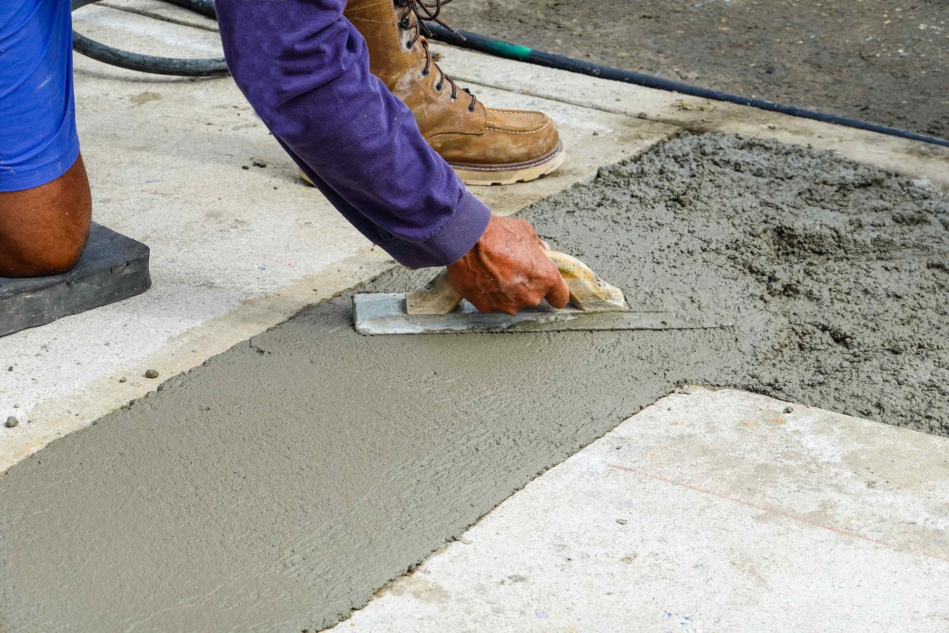 A person in a purple shirt uses a metal trowel to smooth wet concrete on a construction site.