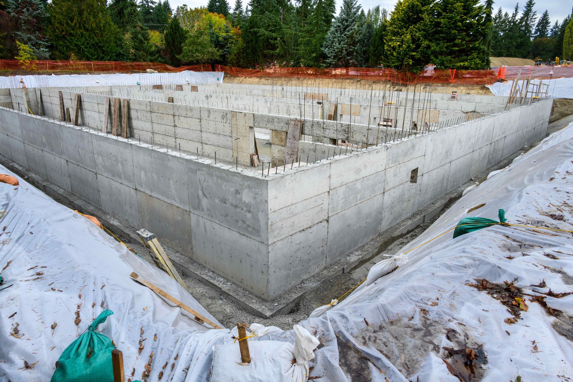 Concrete foundation walls of a building under construction, surrounded by white protective sheeting and forest trees.