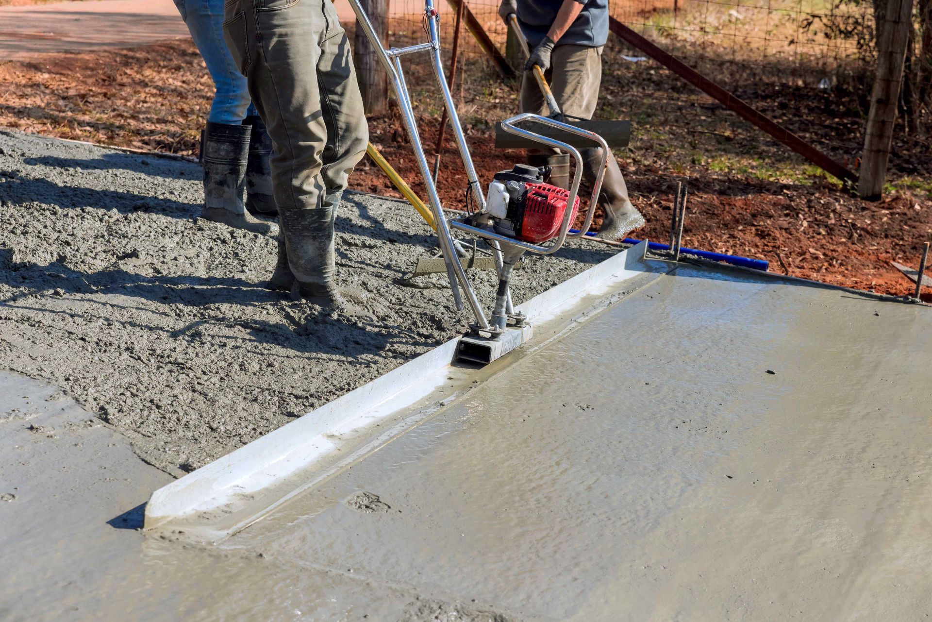 Workers operate a motorized concrete screed to level wet concrete on an outdoor construction site.