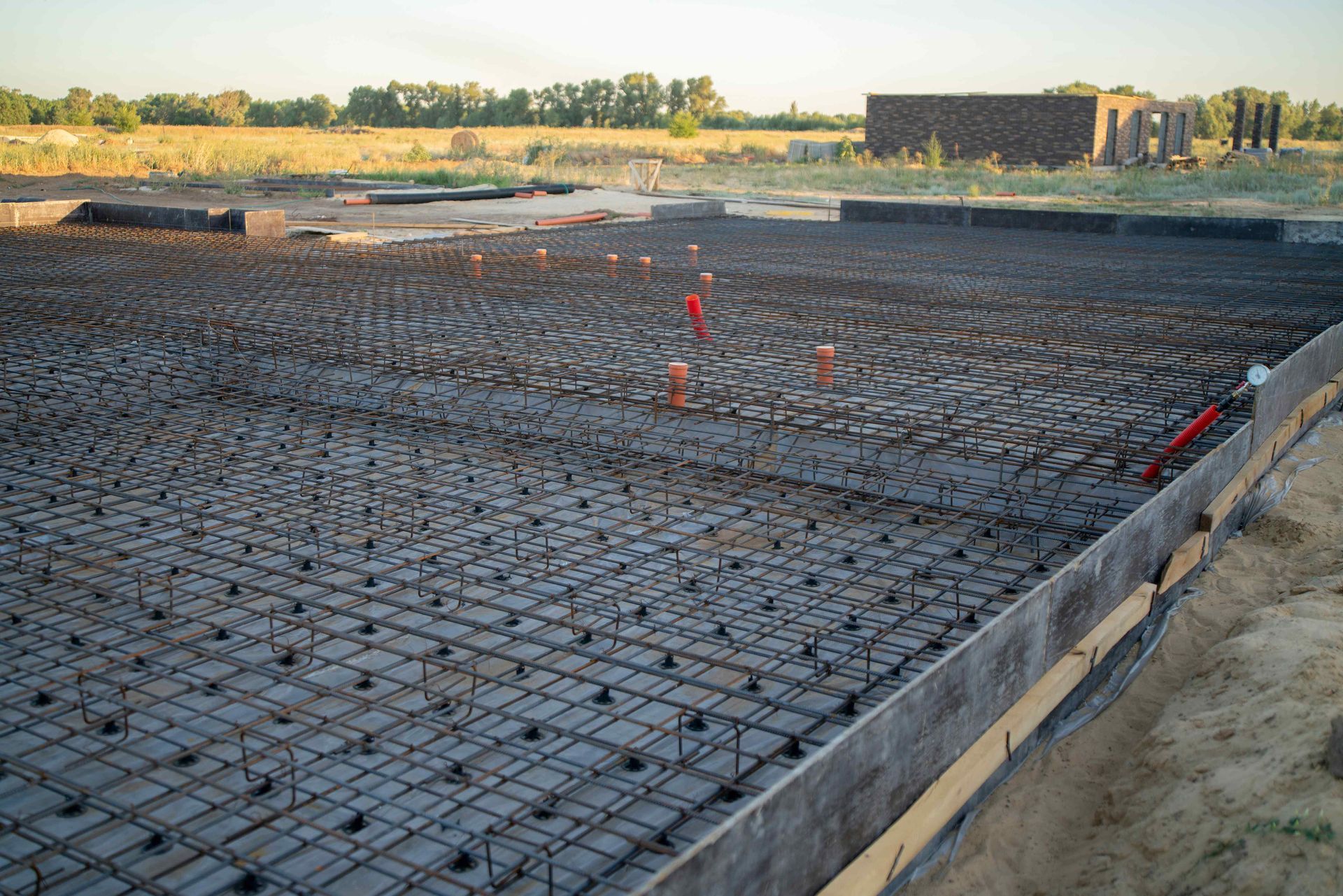 Steel rebar grid laid over a dark plastic vapor barrier for a concrete building foundation in an open field.