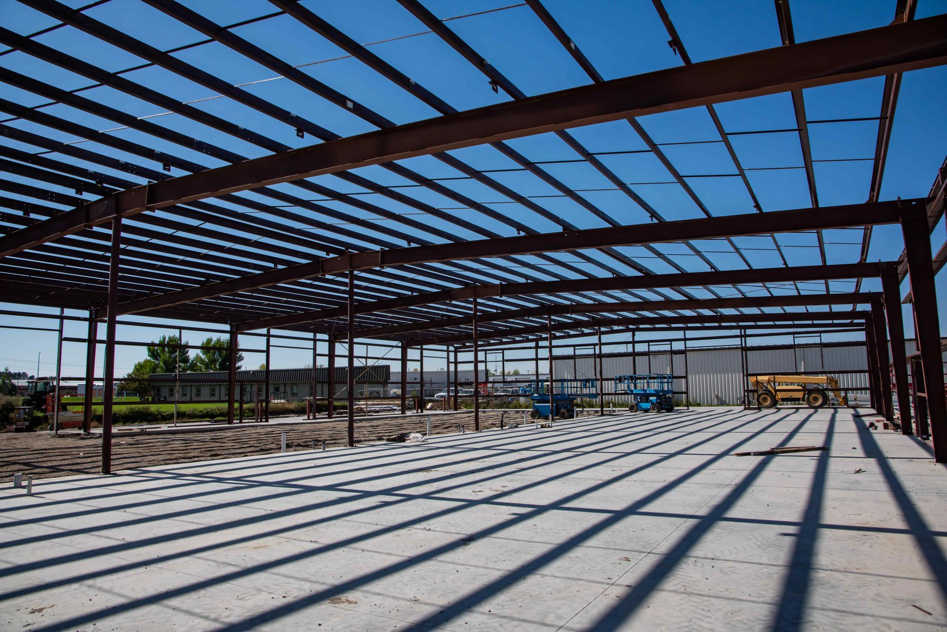 Steel framework and roof beams of a building under construction, casting long parallel shadows on a concrete floor.
