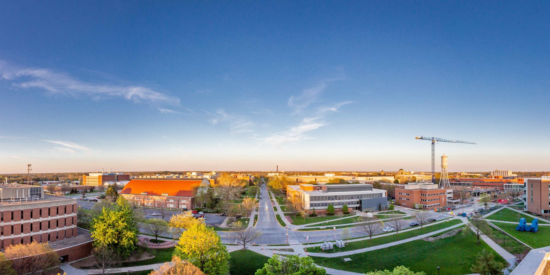 A wide-angle aerial view of a campus or town during golden hour, featuring low-rise buildings, green spaces, and a crane.