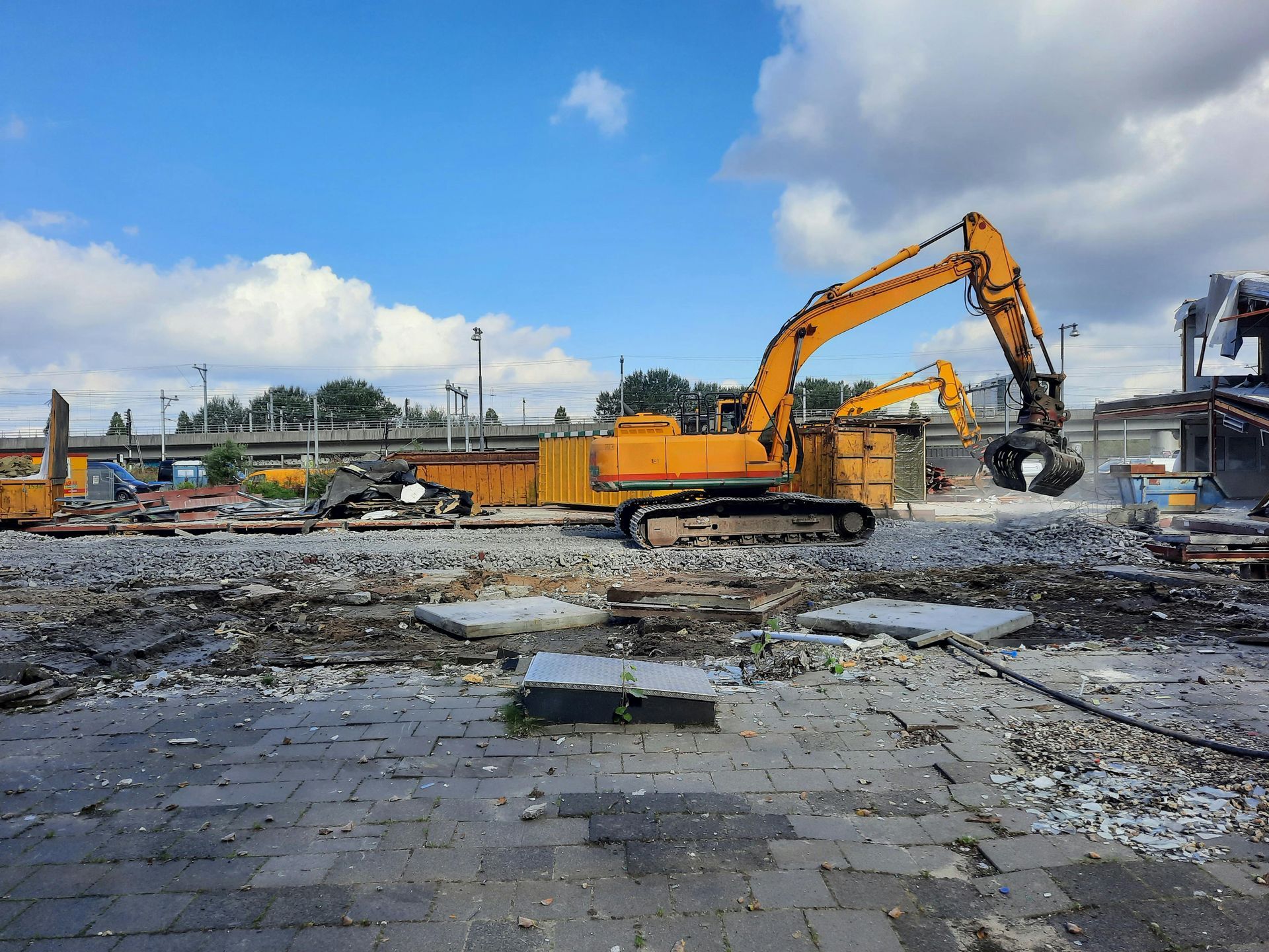 A yellow excavator with a claw attachment operates on a demolition site under a blue, cloudy sky.