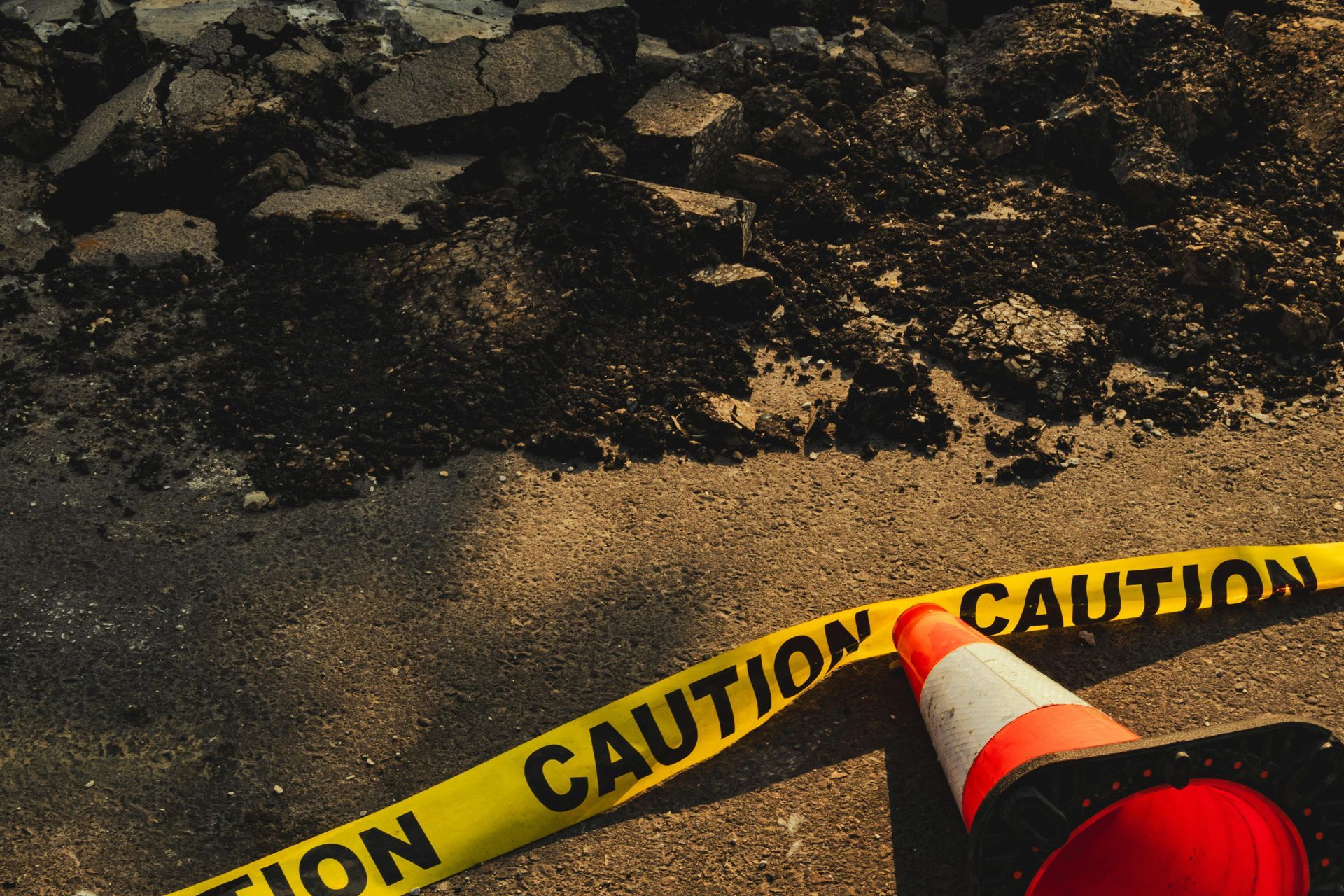 A bright orange traffic cone and yellow caution tape on an asphalt road in front of a section of broken pavement.