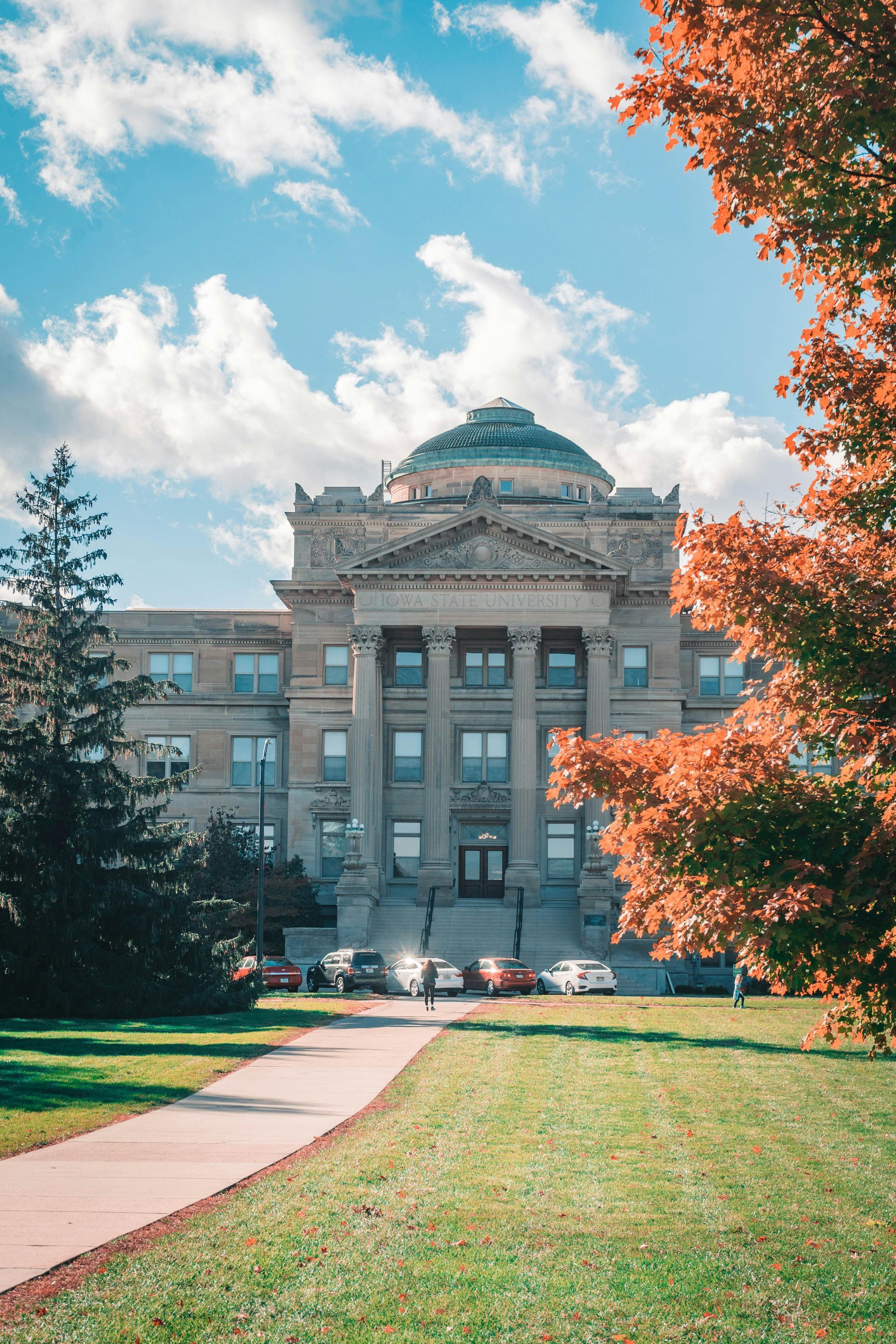 A historic stone building with a dome, seen from a path through a park with fall foliage and green grass under a blue sky.