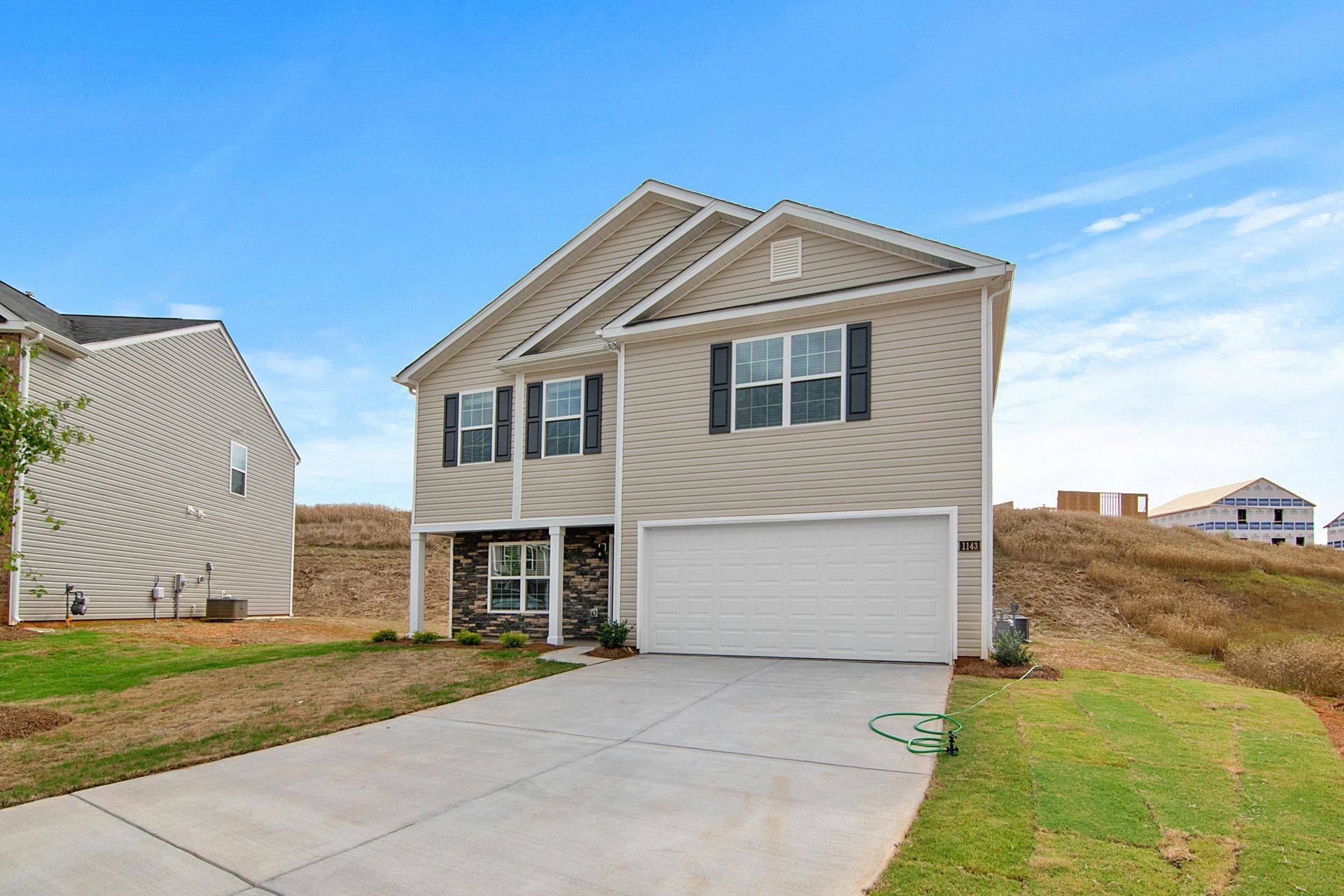 A two-story house with beige siding, a white garage, and stone accents, set on a grassy lot under a clear blue sky.