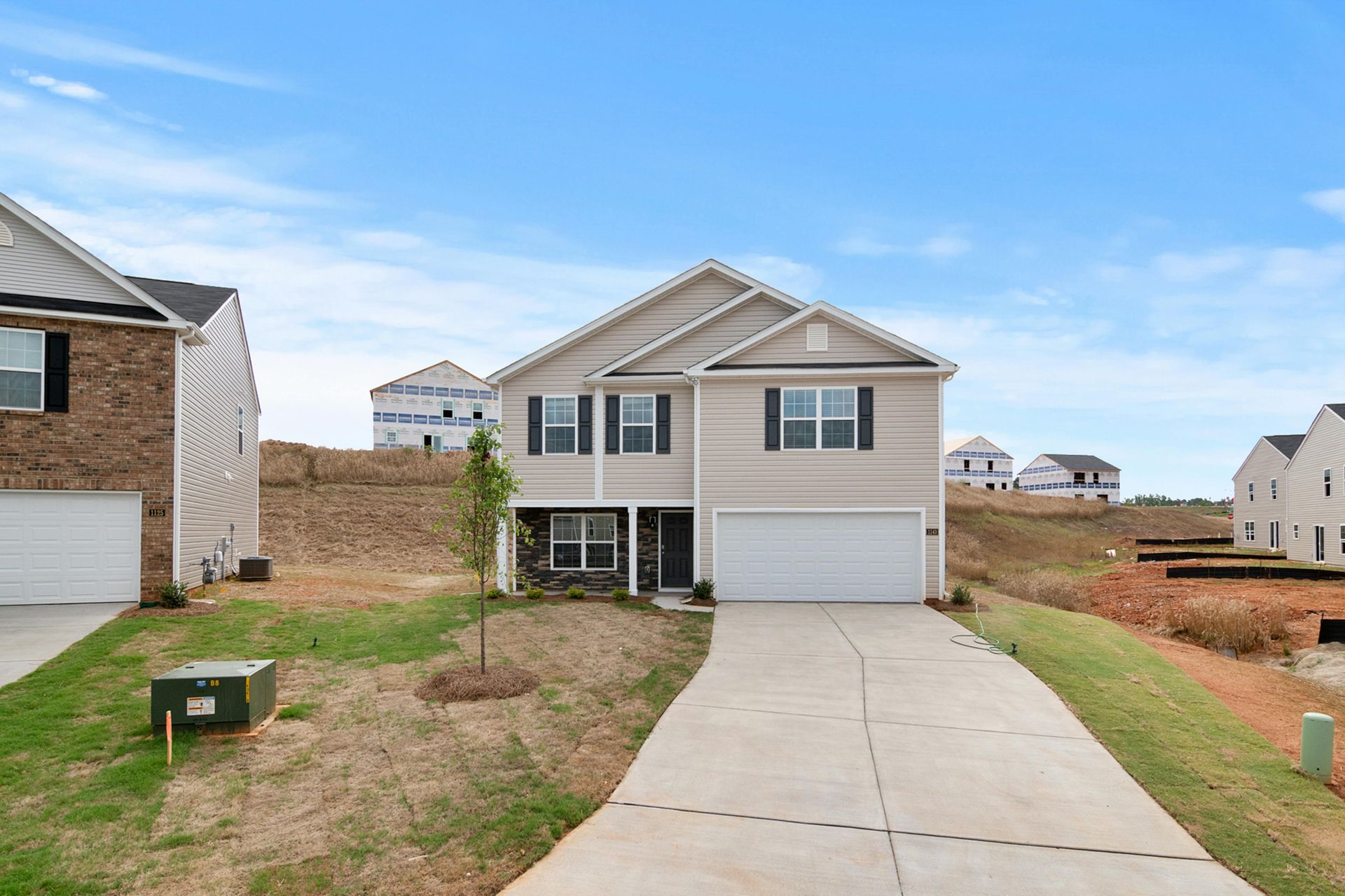 A light-colored two-story suburban house with a white garage, concrete driveway, and grassy yard in a developing neighborhood.