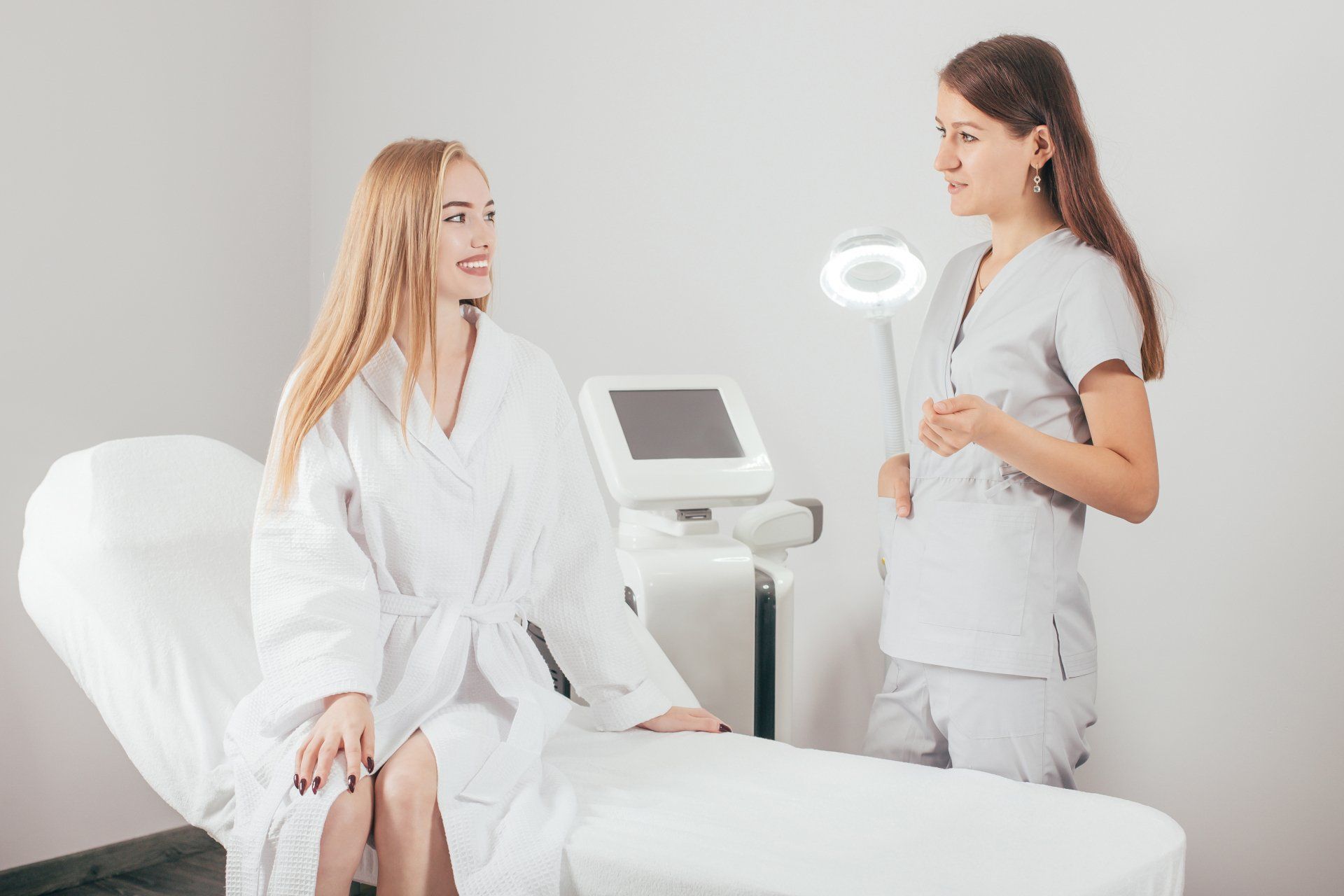 Woman in robe sits on exam table; technician in scrubs talks to her in a clinic.