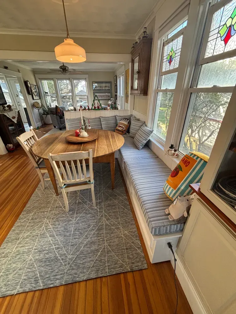 Bright dining room with wooden table, bench seating, patterned rug, and sunlit windows.