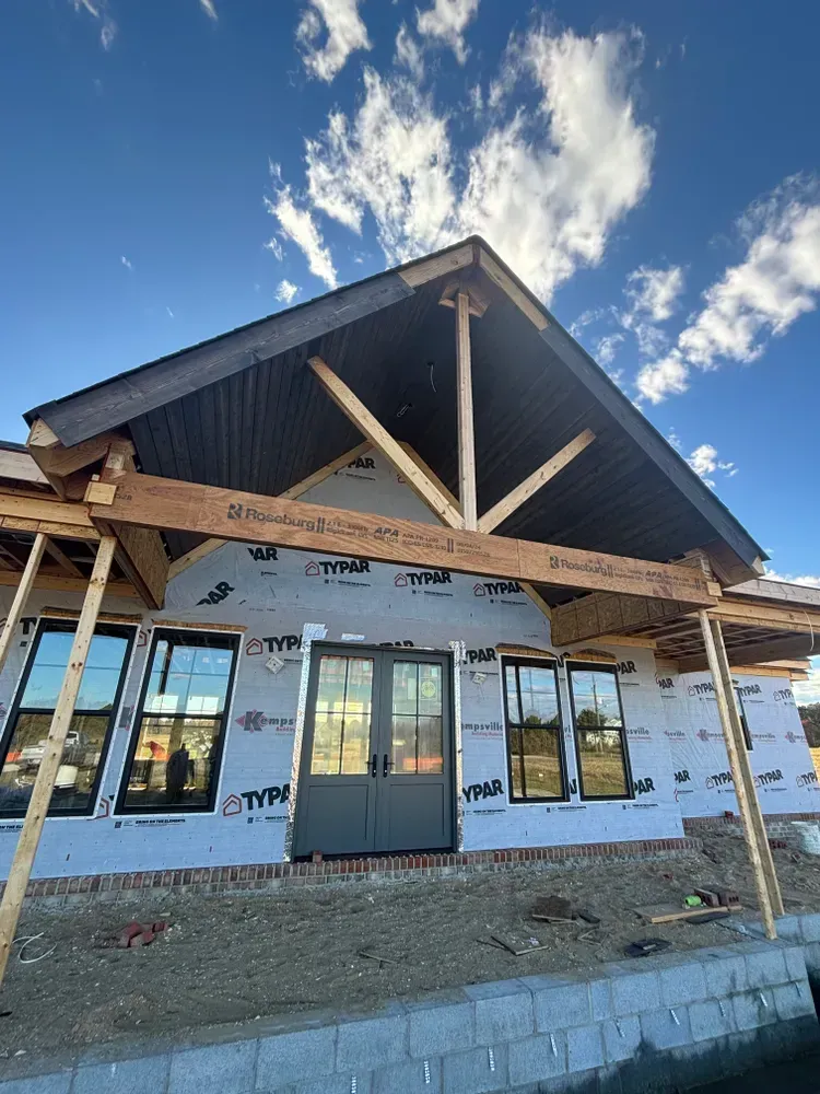 Partially finished house exterior with dark roof, white wrap, and windows under a blue sky