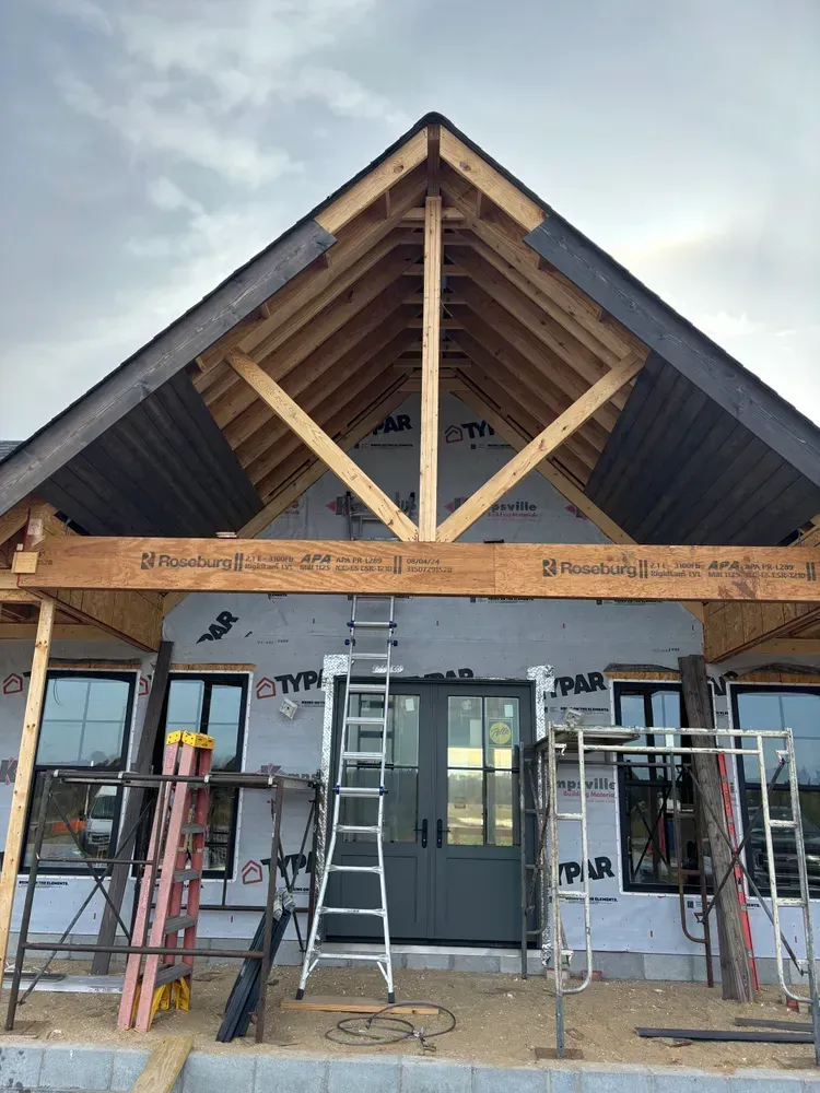 House under construction with exposed roof trusses, front porch framing, ladders, and scaffolding