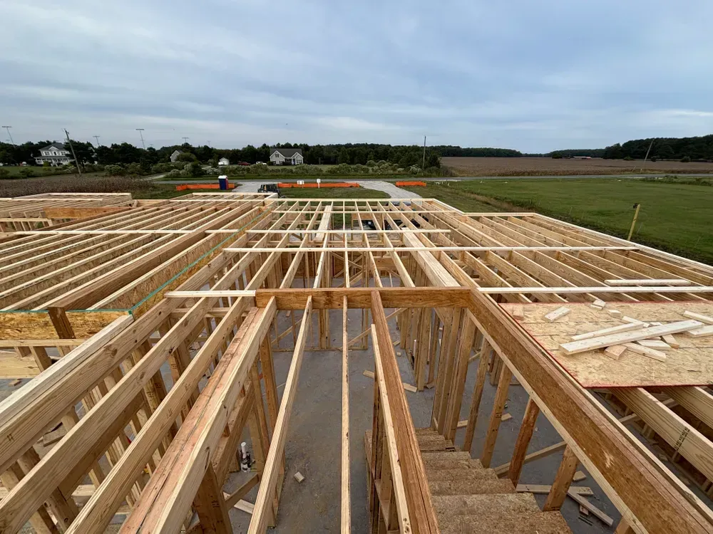 Wooden house framing under construction on a cleared site with open fields in the background