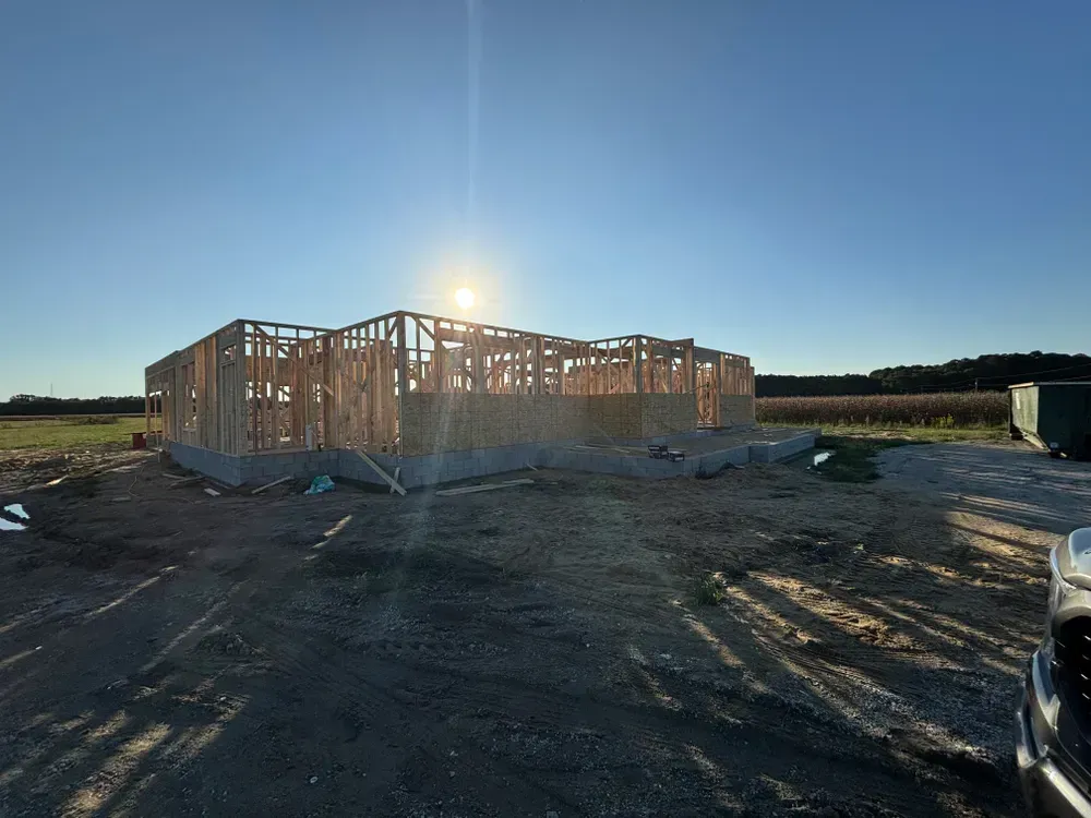 Wood-framed house under construction at sunset on a dirt lot, with a vehicle edge visible in the foreground.