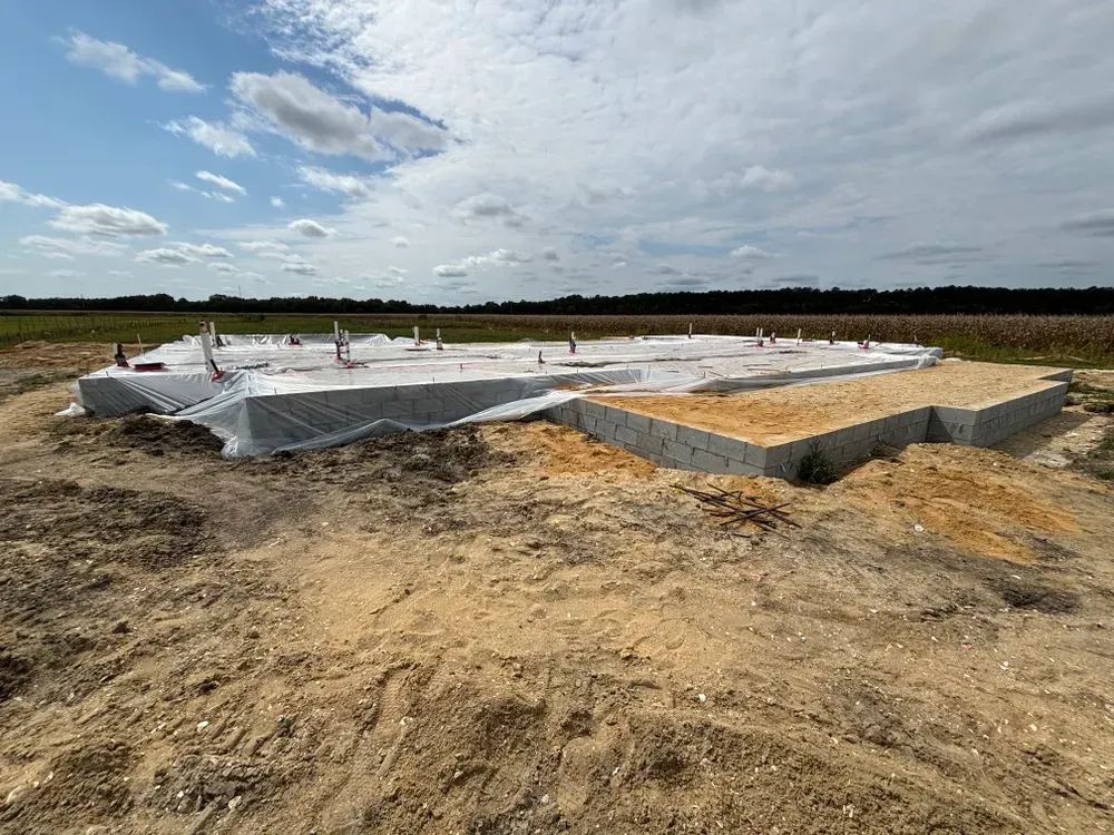 Concrete foundation under construction on a dirt lot with wrapped building materials and cloudy sky
