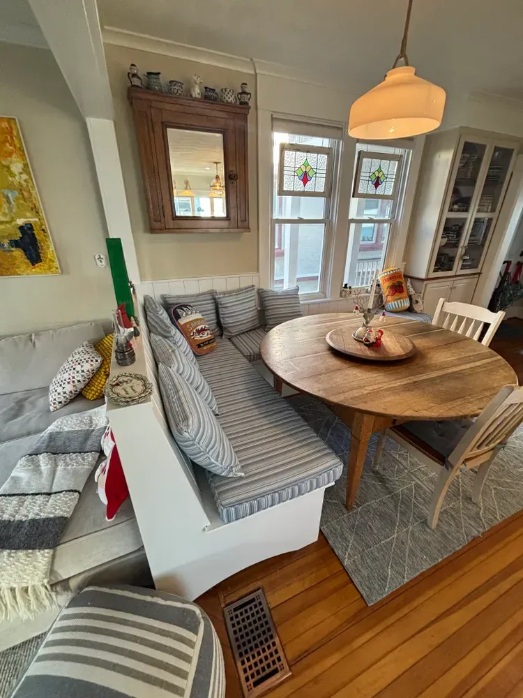 Cozy dining nook with round wooden table, striped built-in bench, chairs, and pendant light by windows