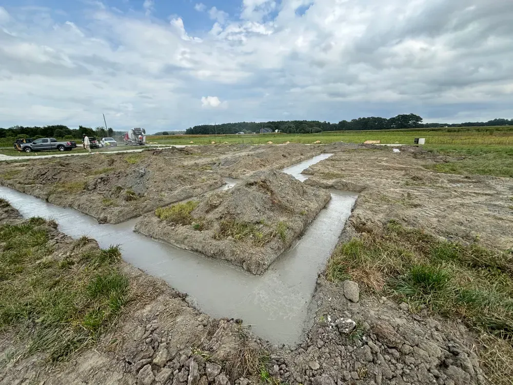 Excavation trench network in a muddy field under a cloudy sky, with vehicles in the distance.