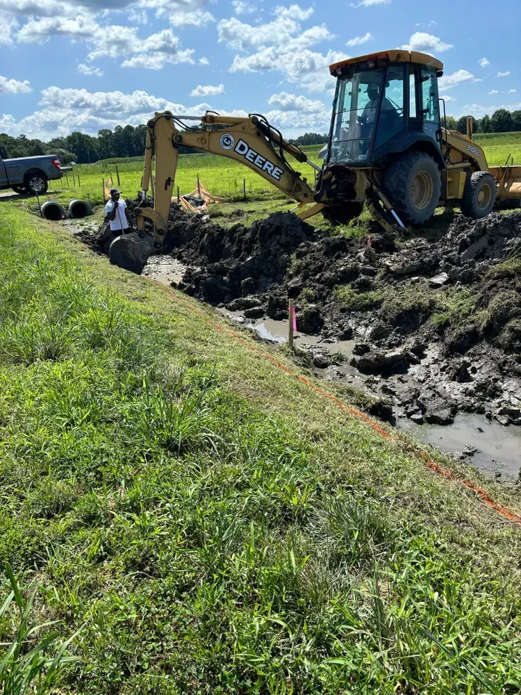 Yellow backhoe digging muddy ditch beside grassy field under a blue sky