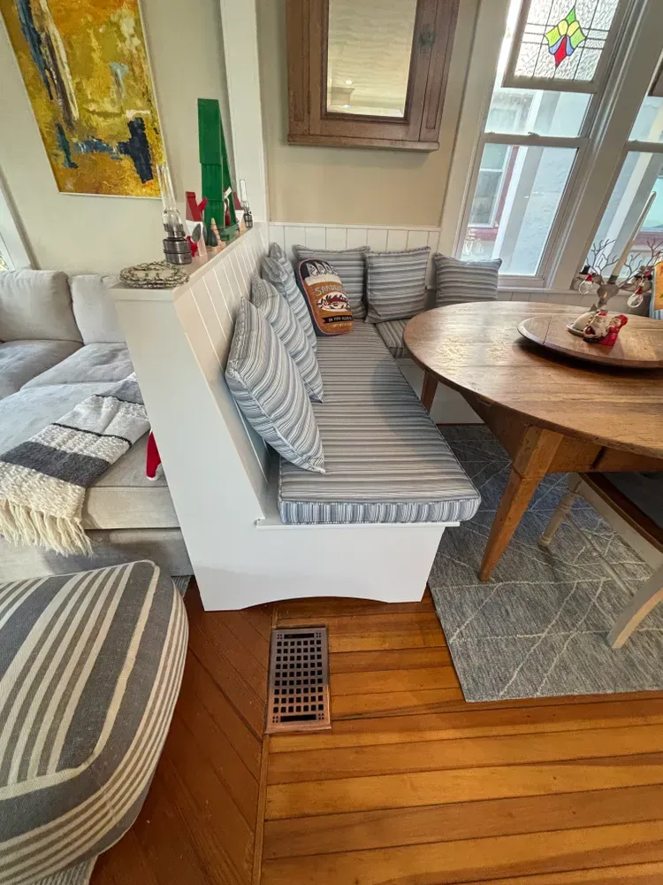 Cozy sunroom with striped couches, round wooden table, and hardwood floor by a window and small vent.