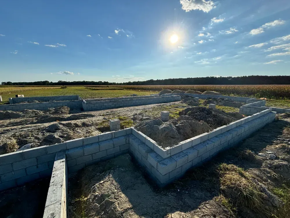 Concrete foundation walls at a rural construction site under a bright sun, with fields and trees in the background