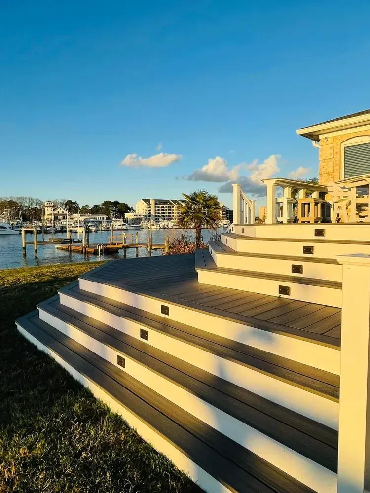 Sunlit waterfront steps beside a dock and columns, with blue sky and boats in the distance