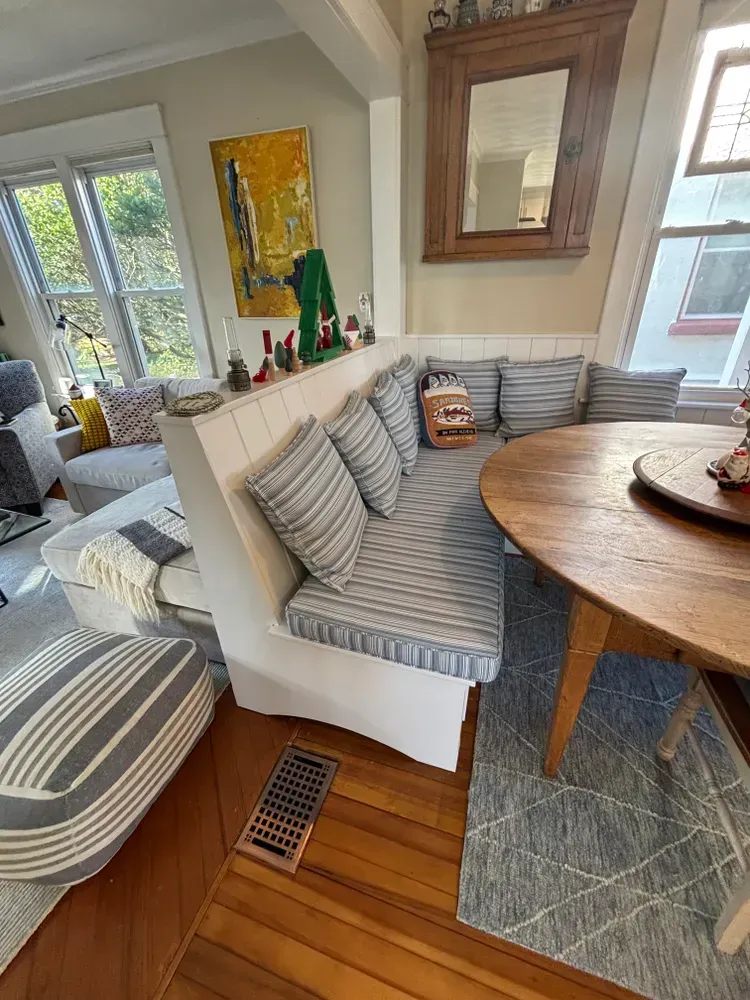 Bright sunroom with striped built-in bench, round wooden table, rug, windows, and wall art