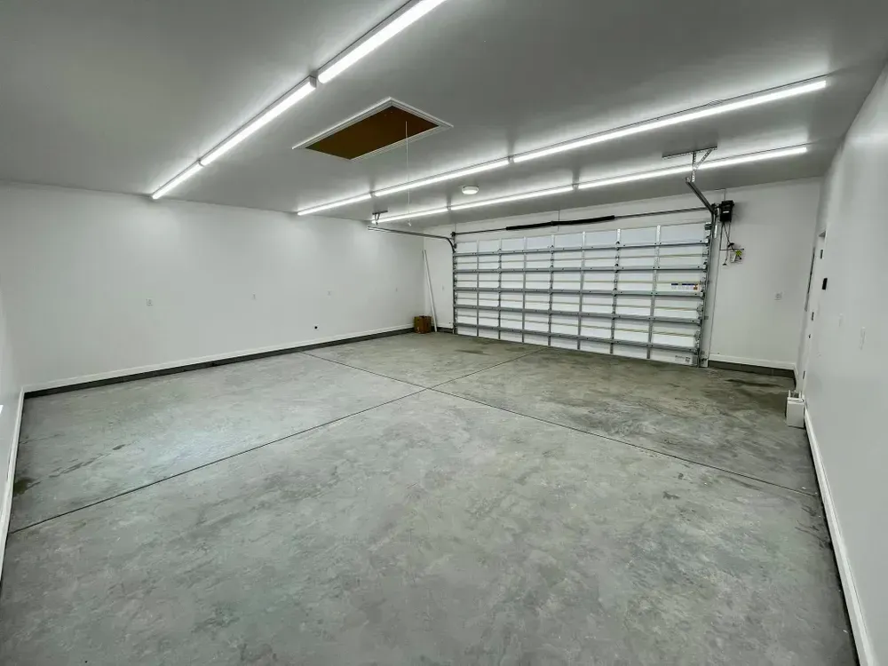 Empty garage with concrete floor, white walls, fluorescent lights, and a closed roll-up door.