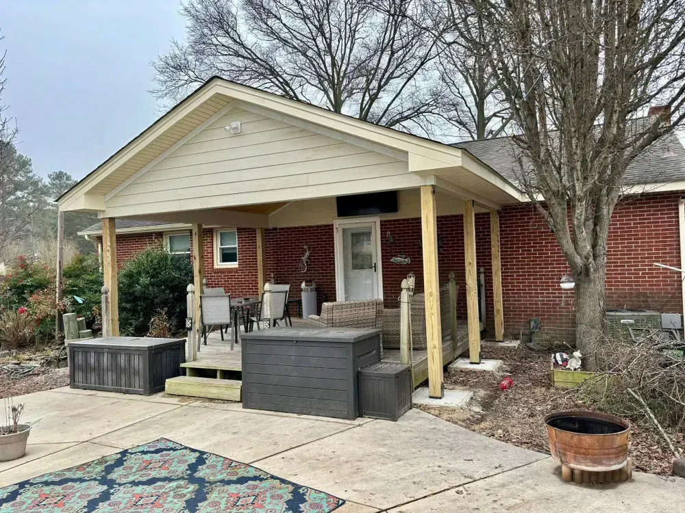 Small brick house porch with columns, chairs, potted plants, and a concrete patio under bare trees.