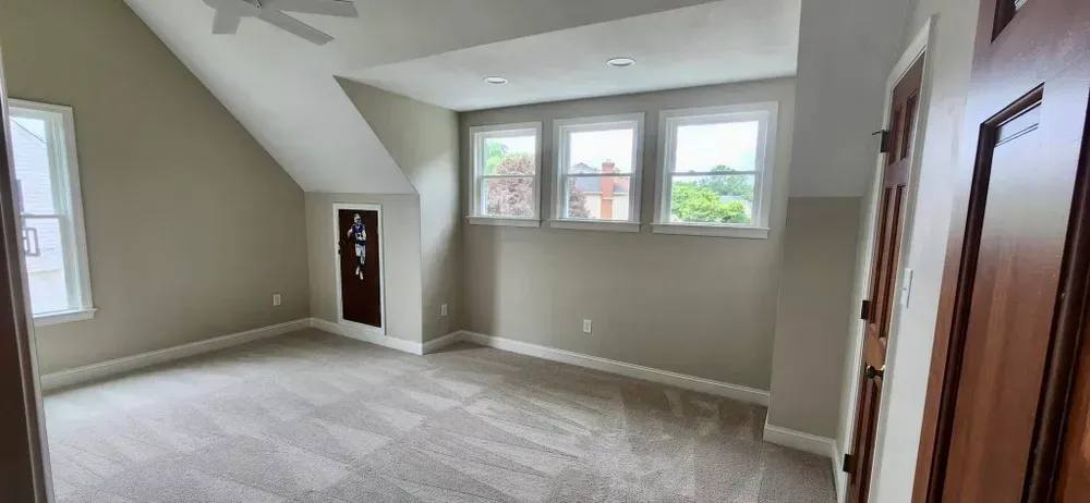 Spacious carpeted room with sloped ceiling, three windows, gray walls, and wood-trimmed doors