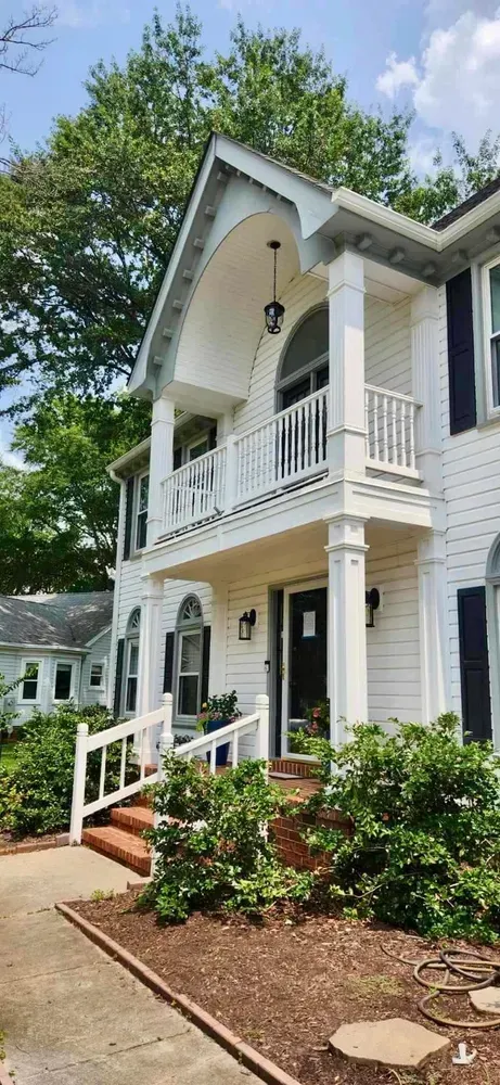 White two-story house with a front porch, black shutters, and steps, surrounded by greenery