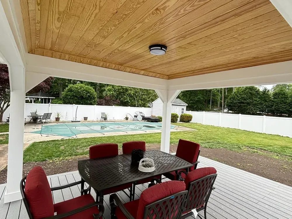 Covered patio dining area beside backyard pool and lawn, with red chairs and a dark table.
