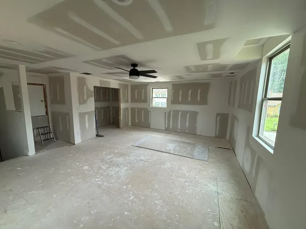Unfinished drywall room with a ceiling fan, windows, and patching compound on the floor and walls.