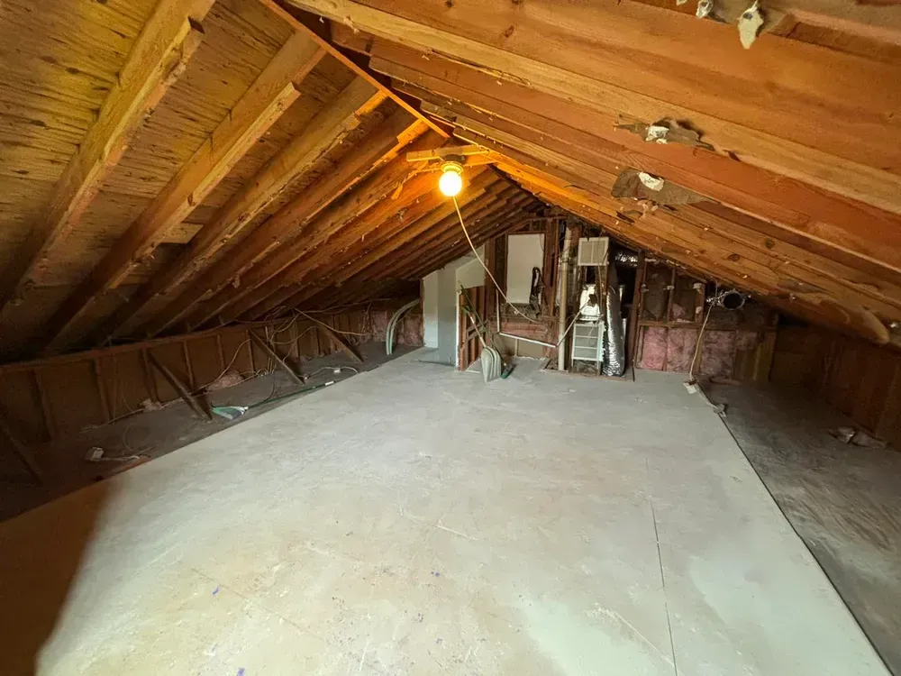 Empty attic with exposed wooden rafters, insulation, and a single hanging light bulb.