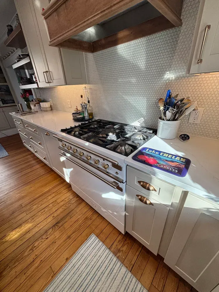 Bright white kitchen with gas range, wood floors, and patterned tile backsplash beneath a wood hood