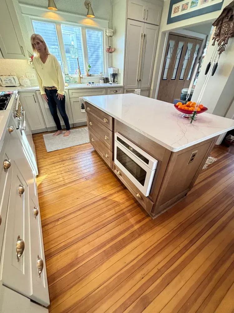 Bright kitchen with wood island, white cabinets, striped floor, and a person standing by the sink under a window