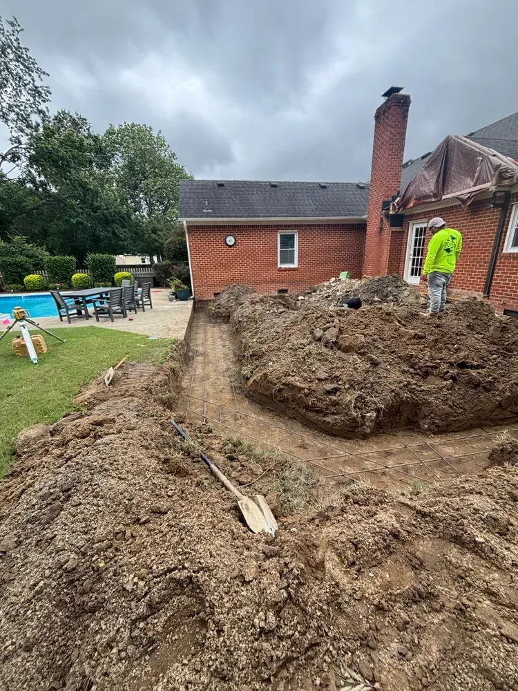 Large muddy excavation pile in a backyard, with a worker in a neon jacket near a brick house.
