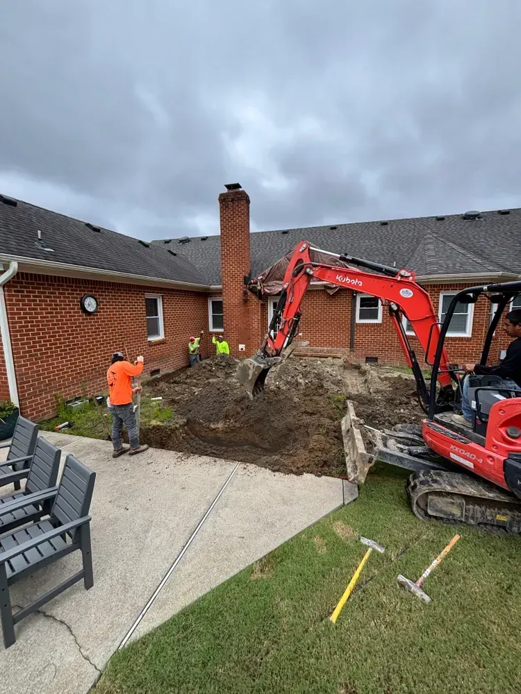 Backyard excavation with red excavator, worker in orange shirt, and brick house under cloudy sky